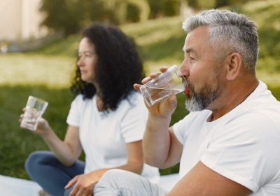 Senior couple is doing yoga outdoors. Stretching in park during sunrise. Brunette in a white t-shirt.